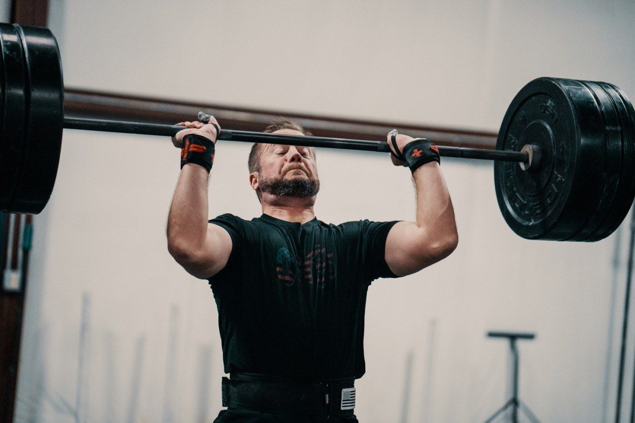 Man lifting a barbell overhead in a gym. Black plates on the bar; he wears a lifting belt and wrist straps.