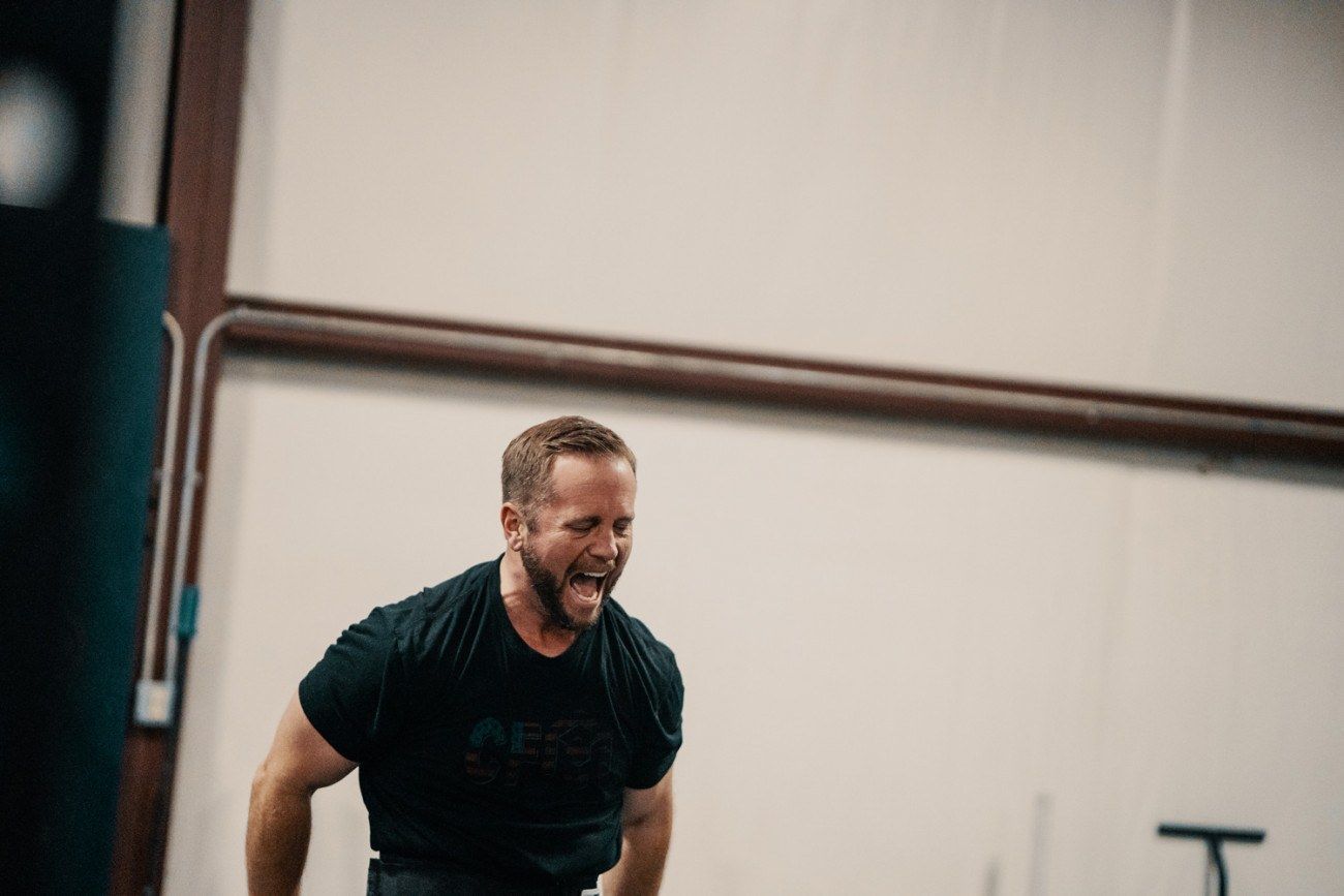 Man in a gym, yelling with exertion. Wearing black shirt, indoors, white walls.