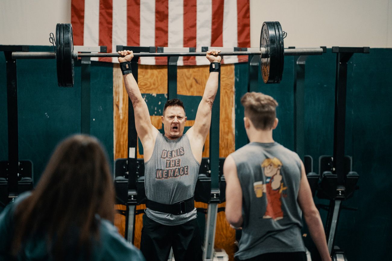 Weightlifter overhead pressing barbell, observed by two people in a gym with American flag.