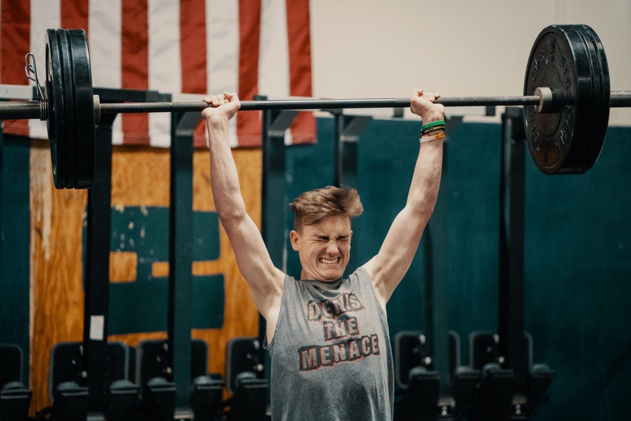 Person lifting a barbell overhead, straining. Indoors with American flag in background.