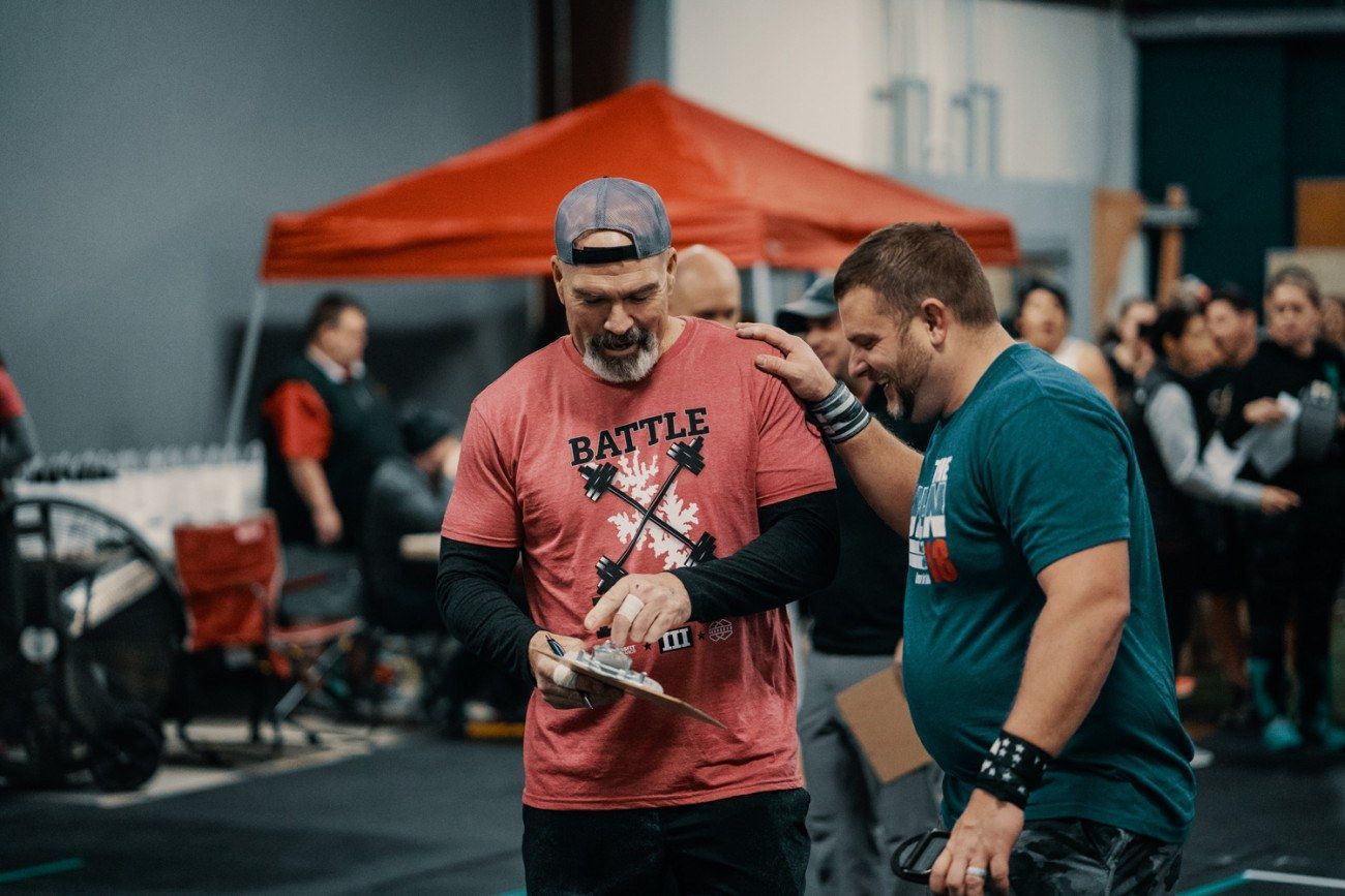 Two men at an athletic event. One in a red shirt reviews a clipboard; other pats his shoulder.