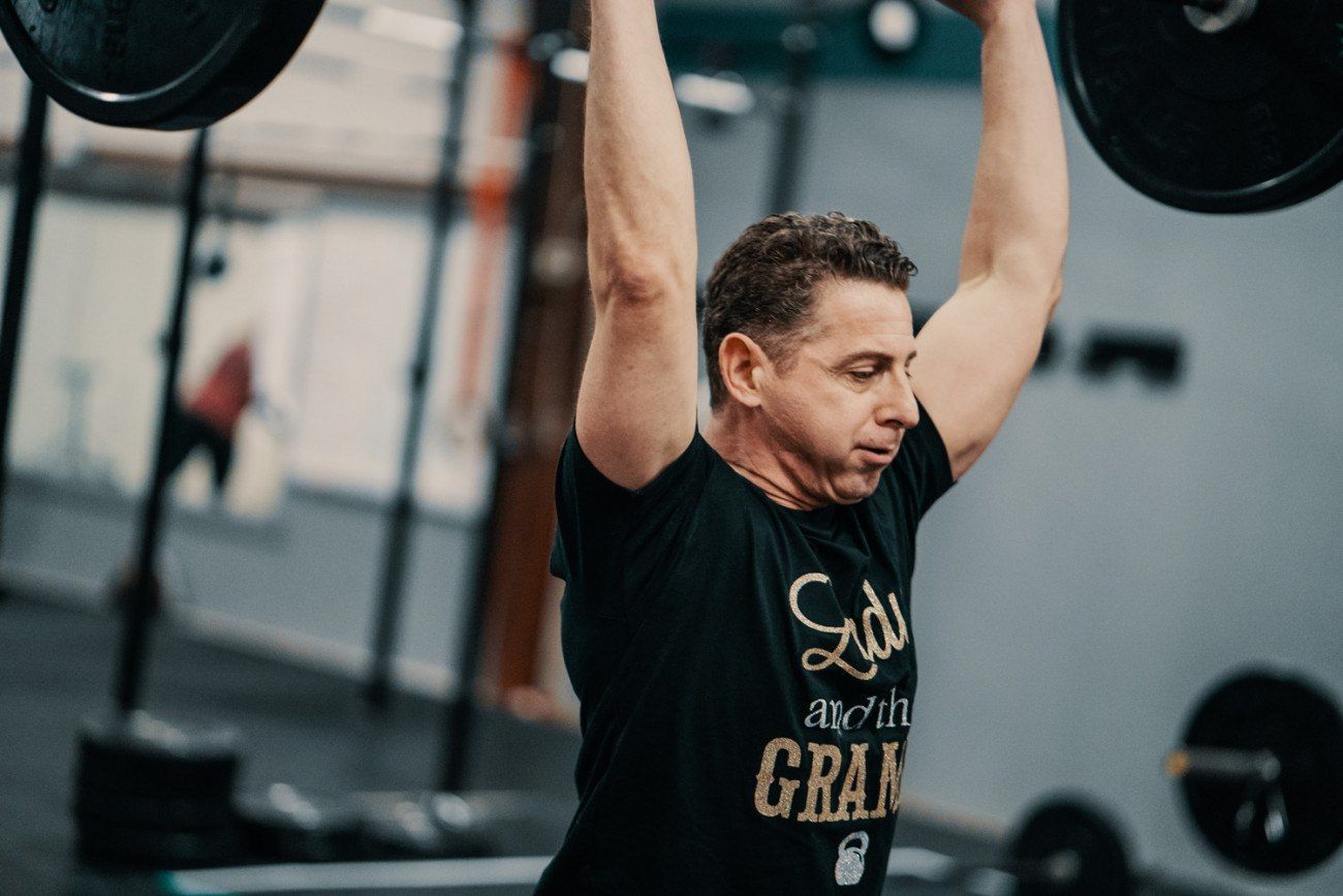 Man lifting barbell overhead in a gym. Black t-shirt, focused expression.