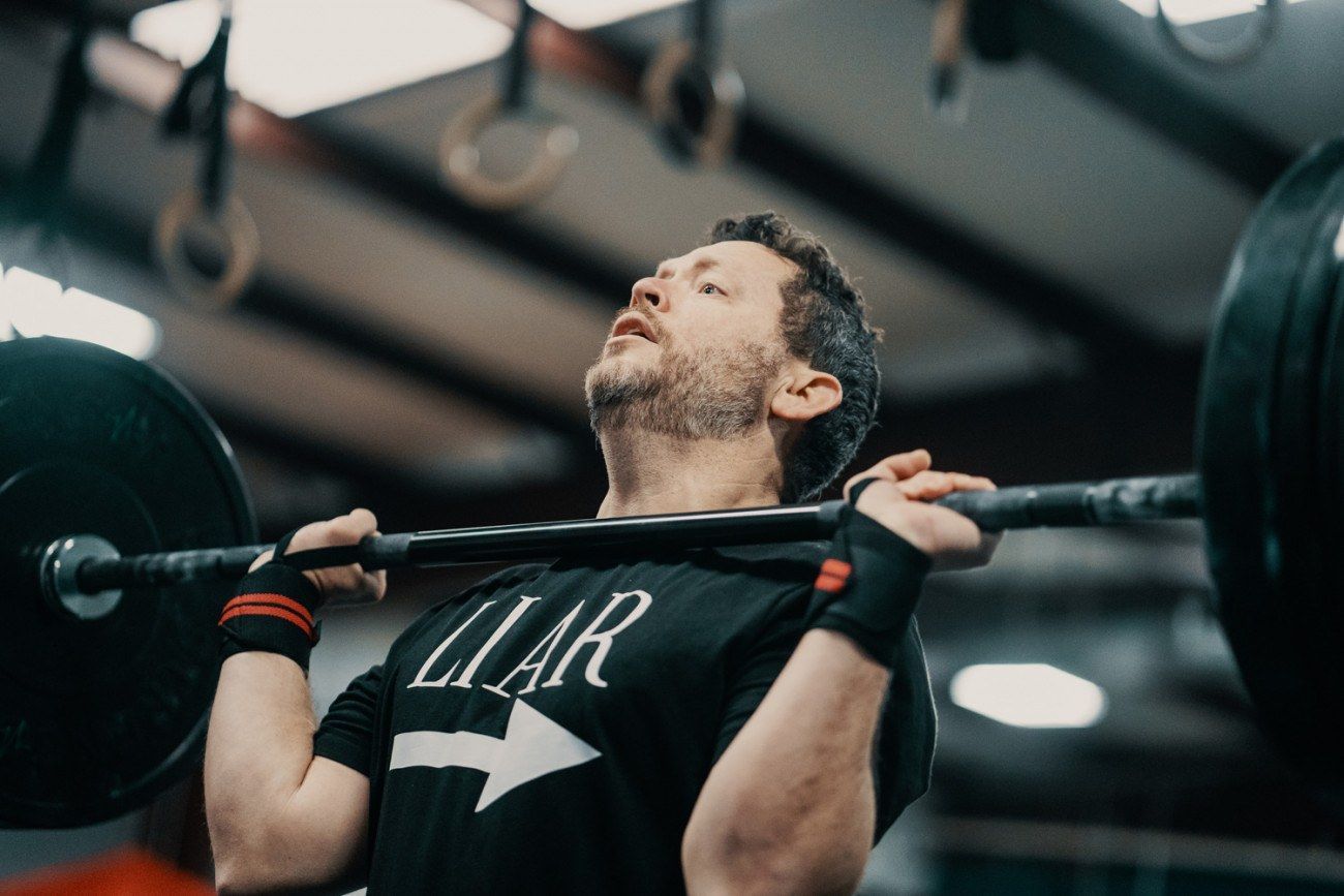 Man lifting barbell overhead in a gym. He is wearing a black shirt with the word 