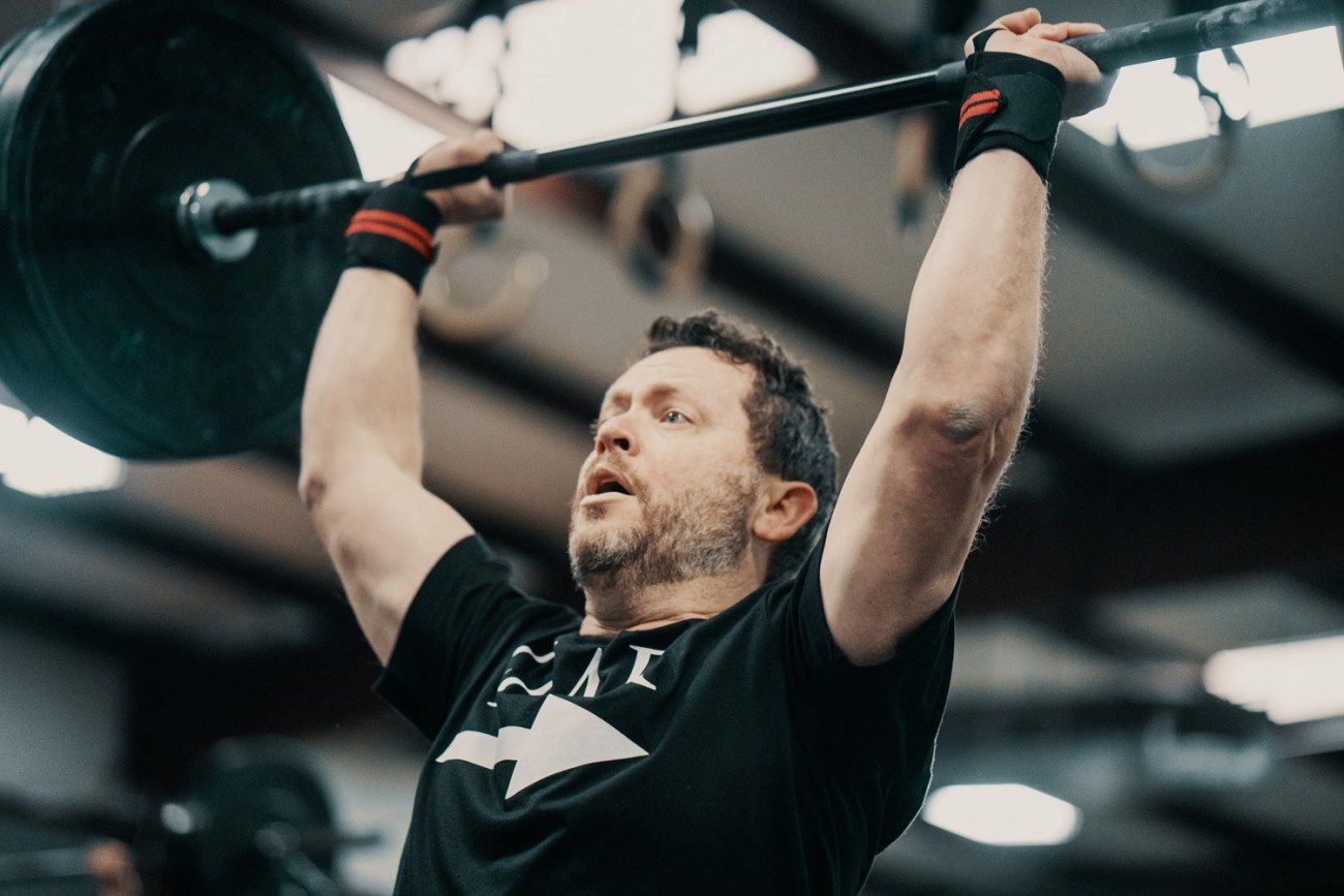 Man lifting barbell overhead in a gym. Black t-shirt, wrist wraps.
