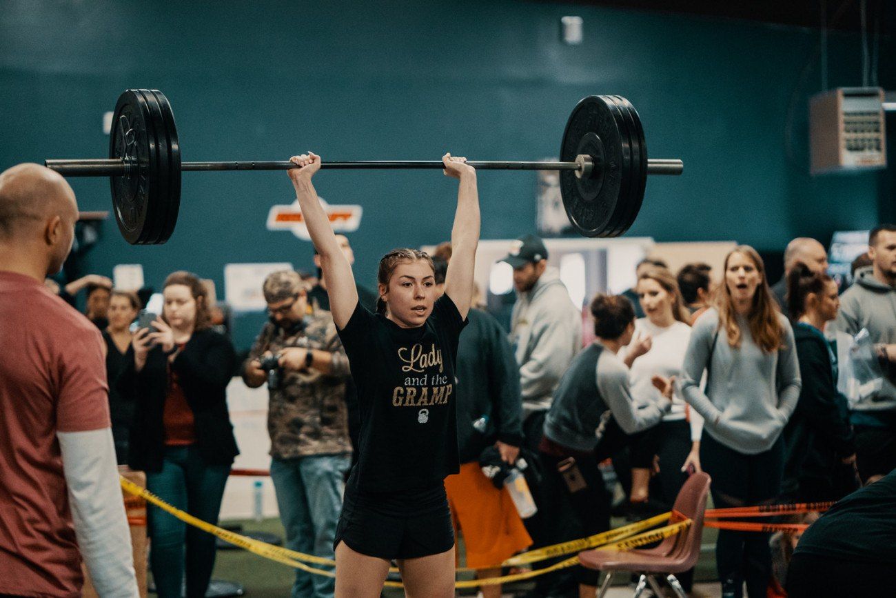 Woman lifts barbell overhead in a gym during competition, arms straight, surrounded by spectators.