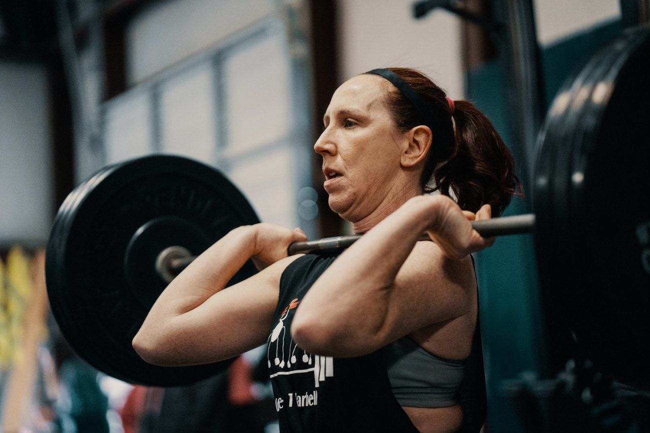 Woman in gym front squatting with a barbell. Exhausted expression, wearing a headband, black tank top.