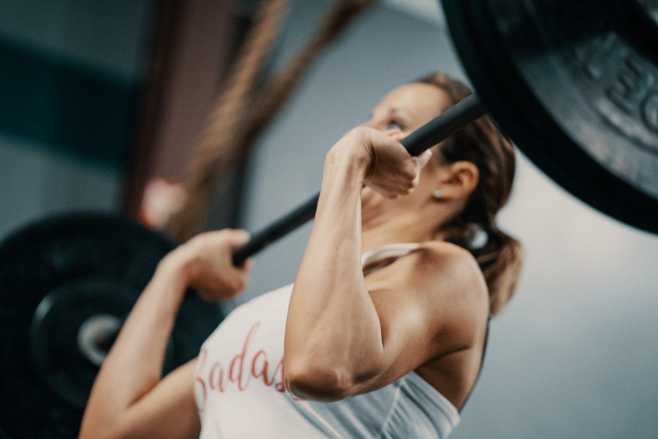 Woman lifting a barbell overhead in a gym. She is focused, wearing a tank top, and the weight is heavy.