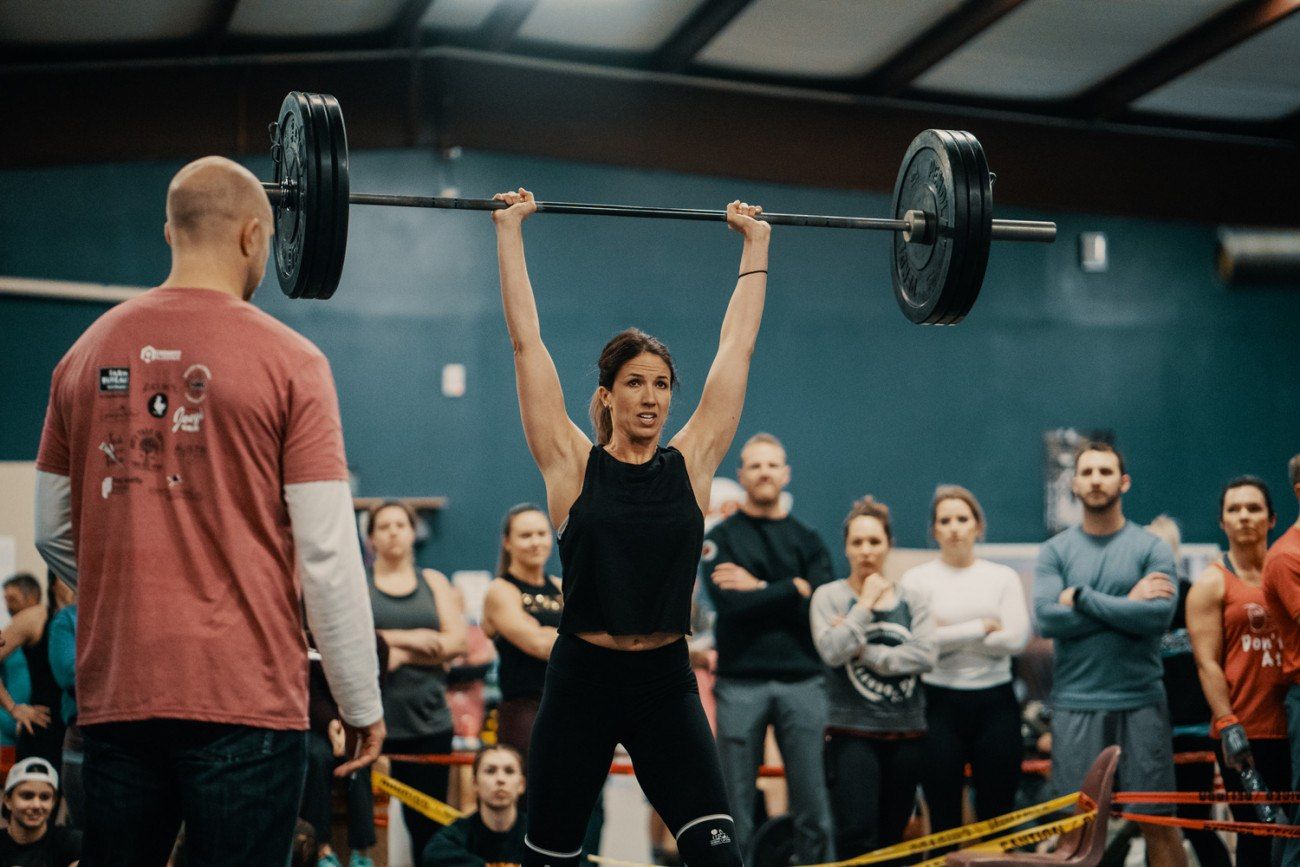 Woman lifting barbell overhead in a gym, watched by a coach and other people.