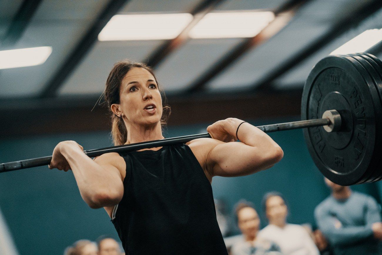 Woman in black top lifting barbell overhead, indoors; focused expression.