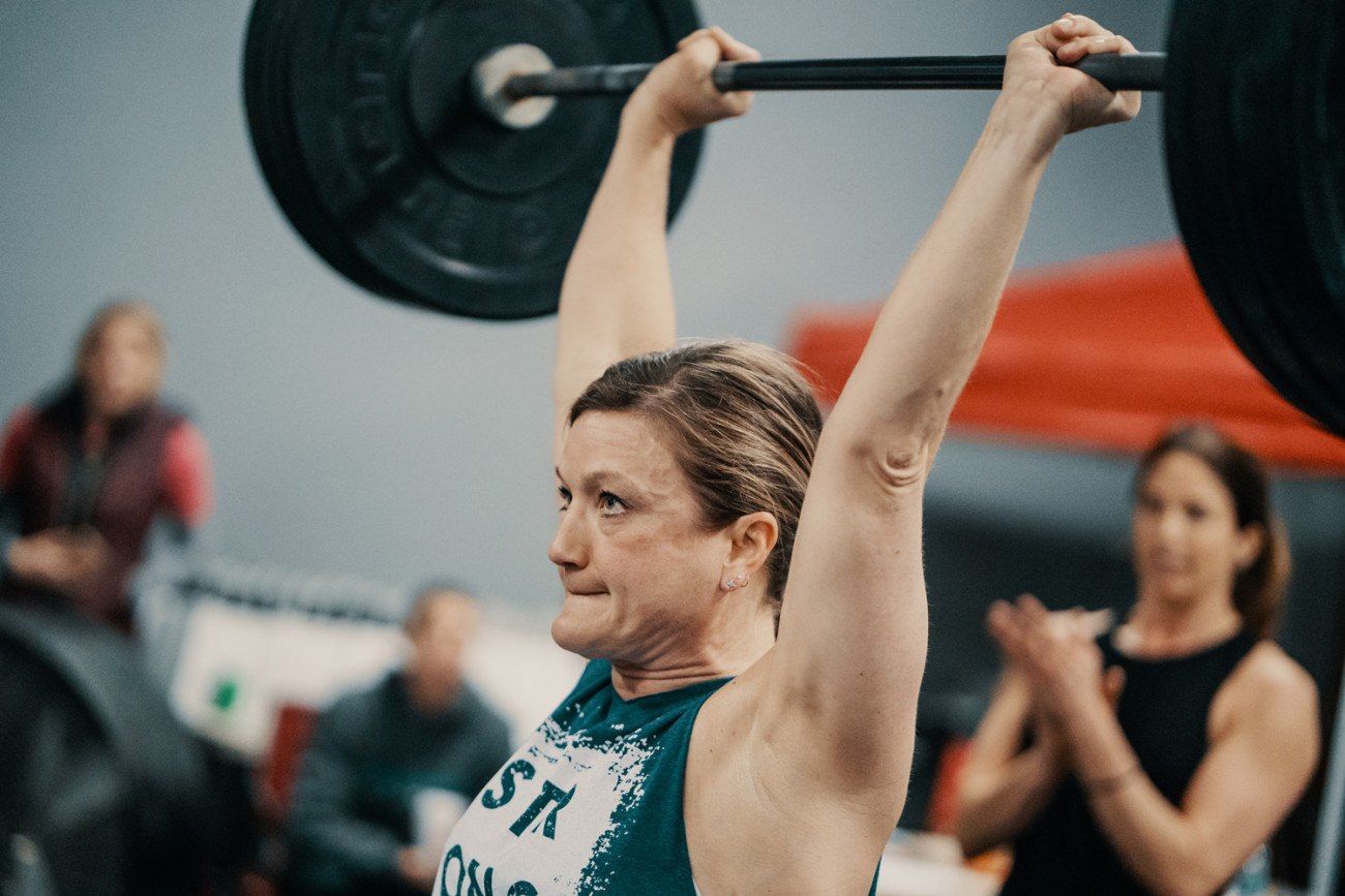 Woman lifting a barbell overhead at a gym, focused expression, onlookers in background, dark plates.