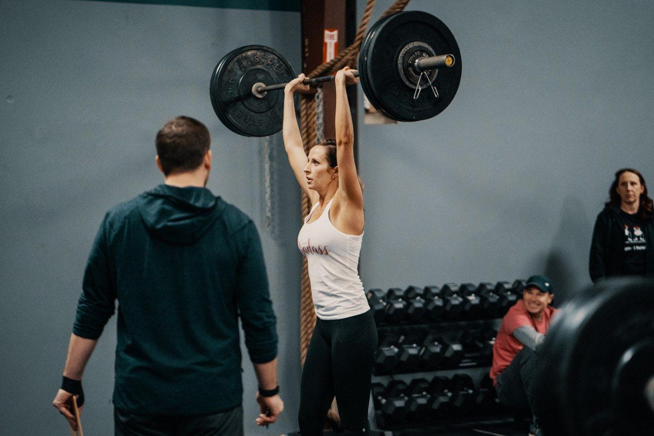 Woman lifting a barbell overhead in a gym, with a spotter watching. Other people are in the background.
