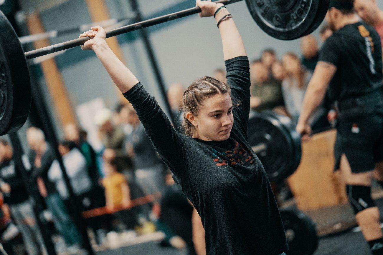 Woman lifts a barbell overhead in a gym, arms extended, crowd in background.
