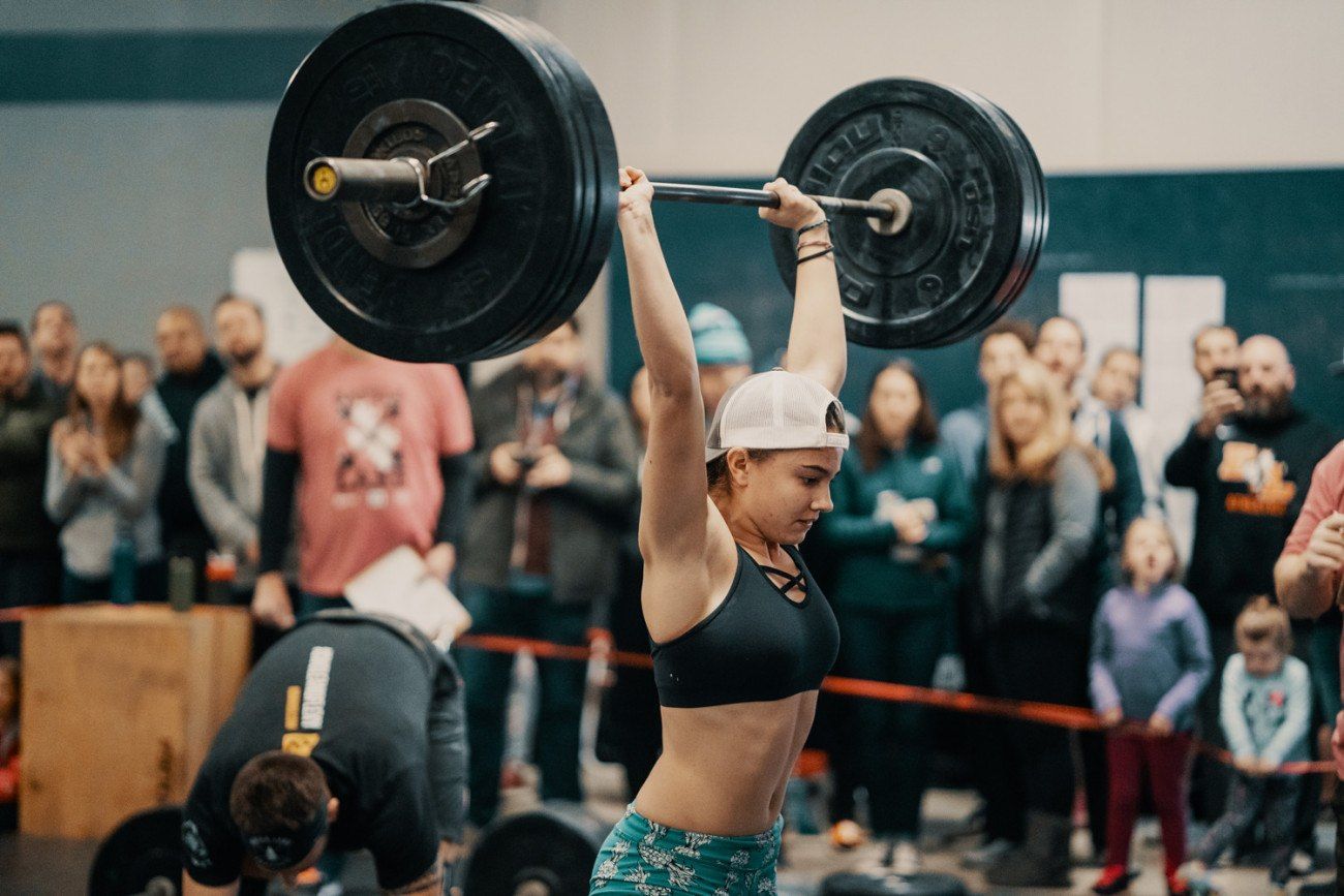 Woman lifting a heavy barbell overhead in a gym, surrounded by spectators.
