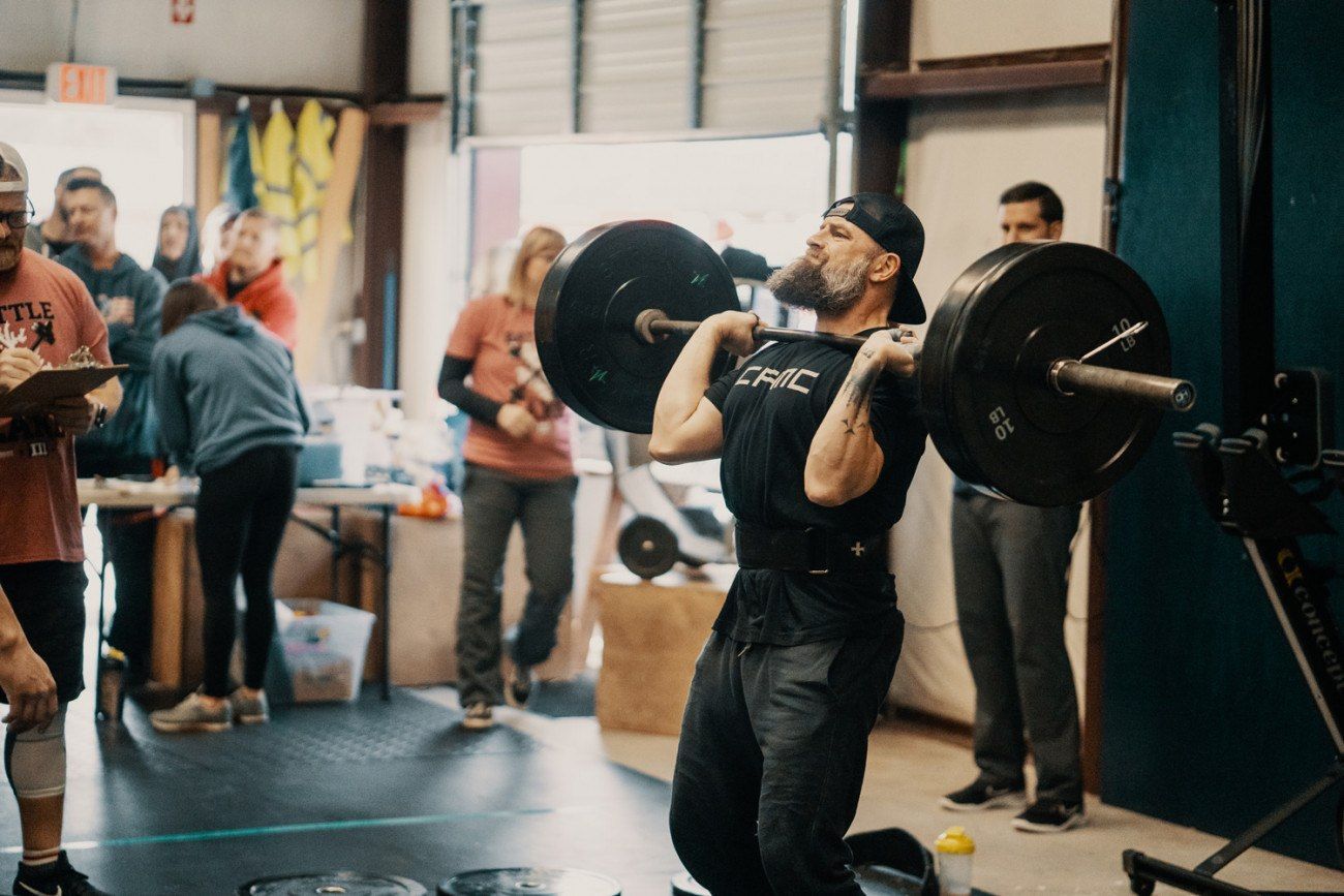 A man in black lifts a barbell overhead in a gym, with onlookers and a judge nearby.