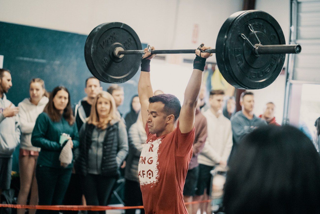 Athlete overhead presses a barbell with onlookers in a gym.
