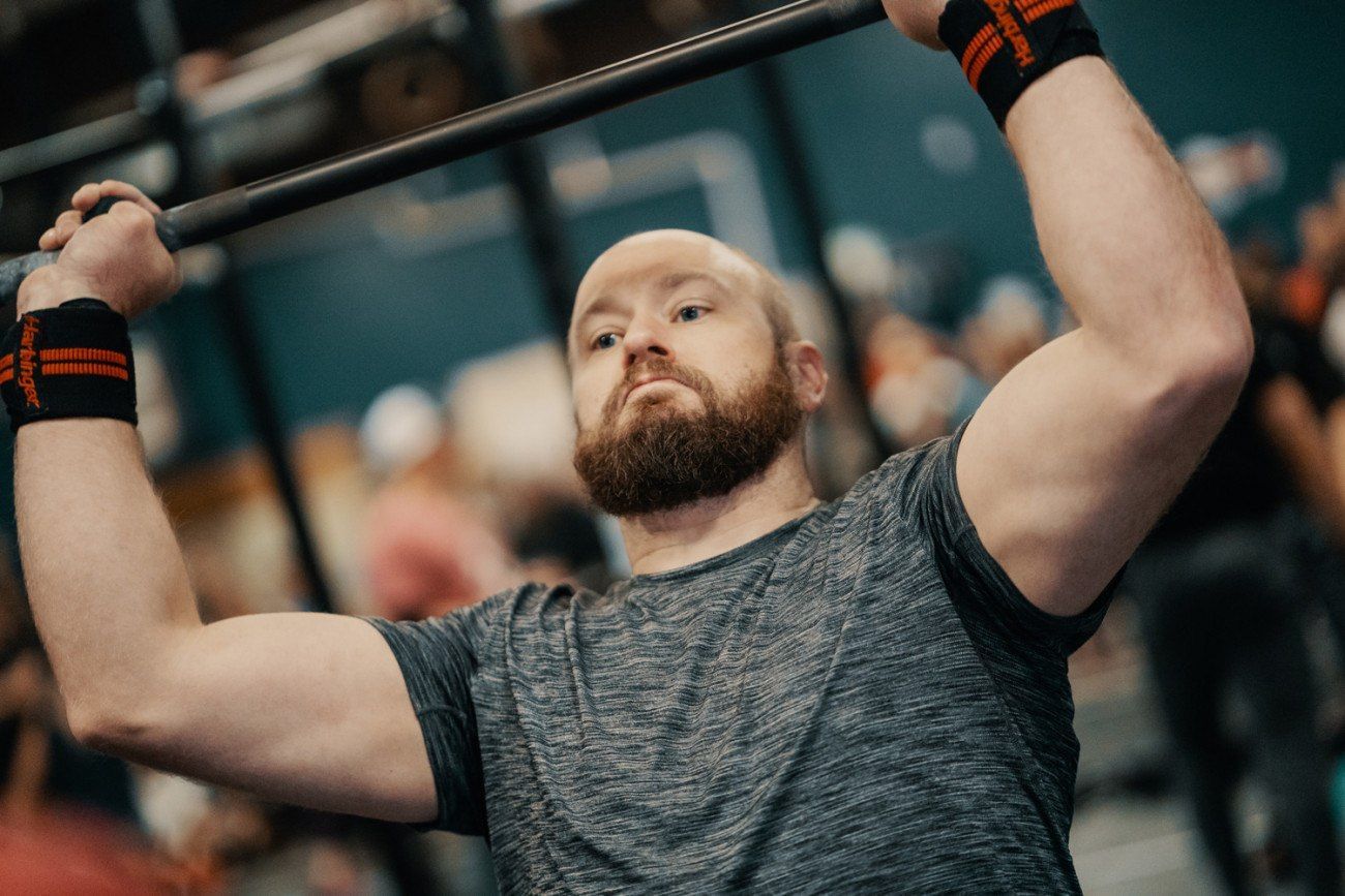 Man lifting a barbell overhead at a gym, wearing wrist wraps.