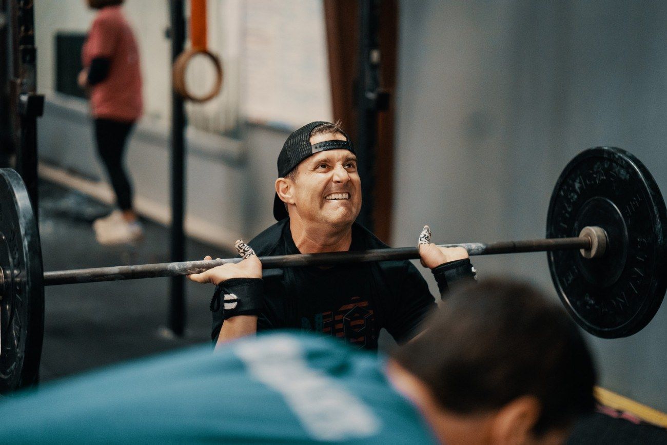 Man in gym, lifting barbell, grimacing. Black hat, shirt, weights. Another person in foreground.