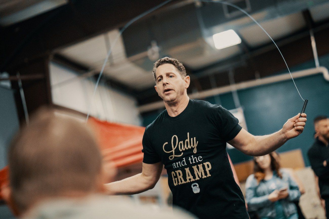 Man in black t-shirt jumps rope indoors. Other people watch.