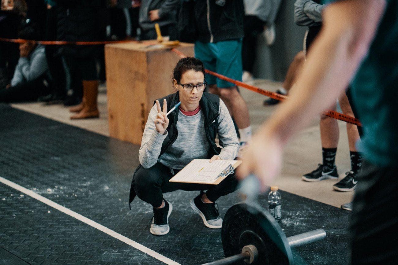 A woman, squatting, watches a weightlifter. She holds a clipboard and pen in a gym setting.