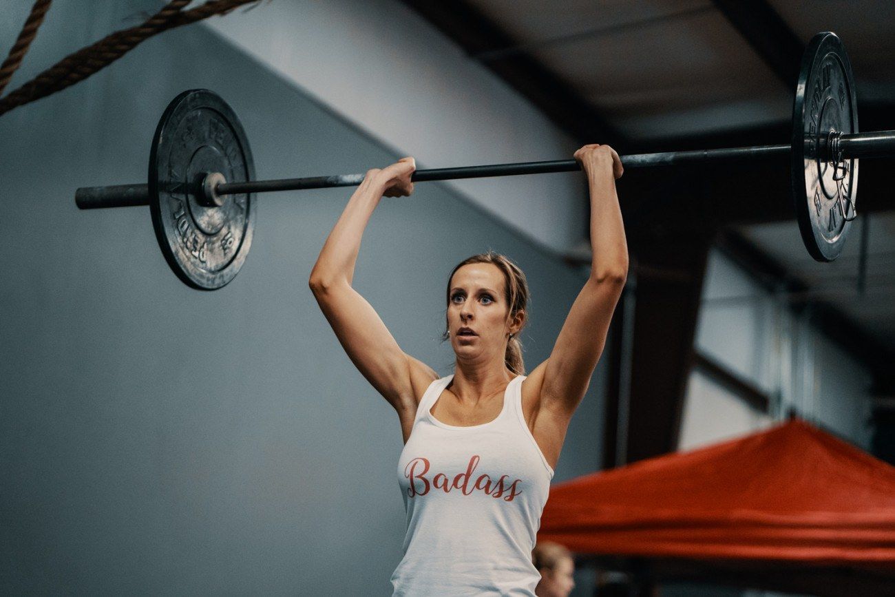 Woman lifting a barbell overhead in a gym. She wears a 