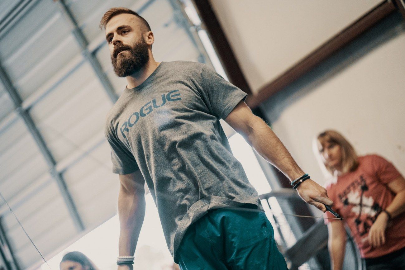 Man with beard in gray Rogue shirt, holding jump rope, in a gym.