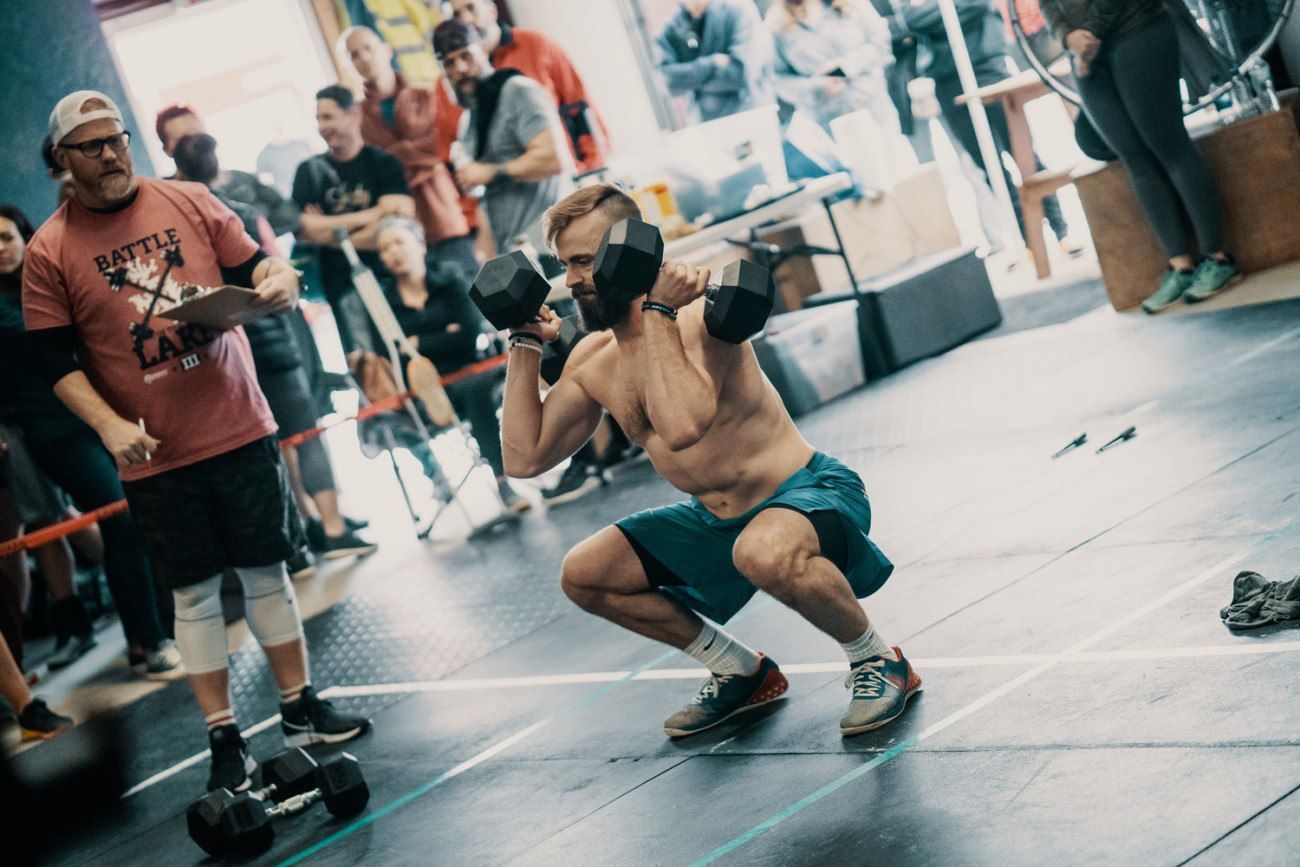 Man doing dumbbell squats in a gym, watched by a judge.