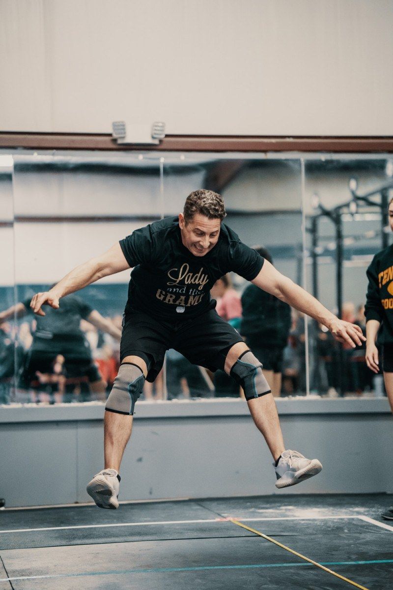 Man wearing knee pads jumps in a gym, arms outstretched, smiling. Reflection in mirror, other people visible.