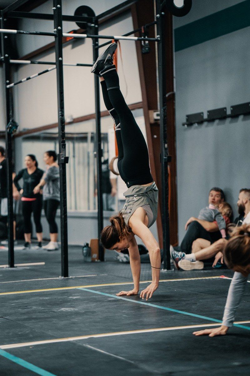 Woman doing a handstand in a gym, wearing leggings and a grey top, others watch.