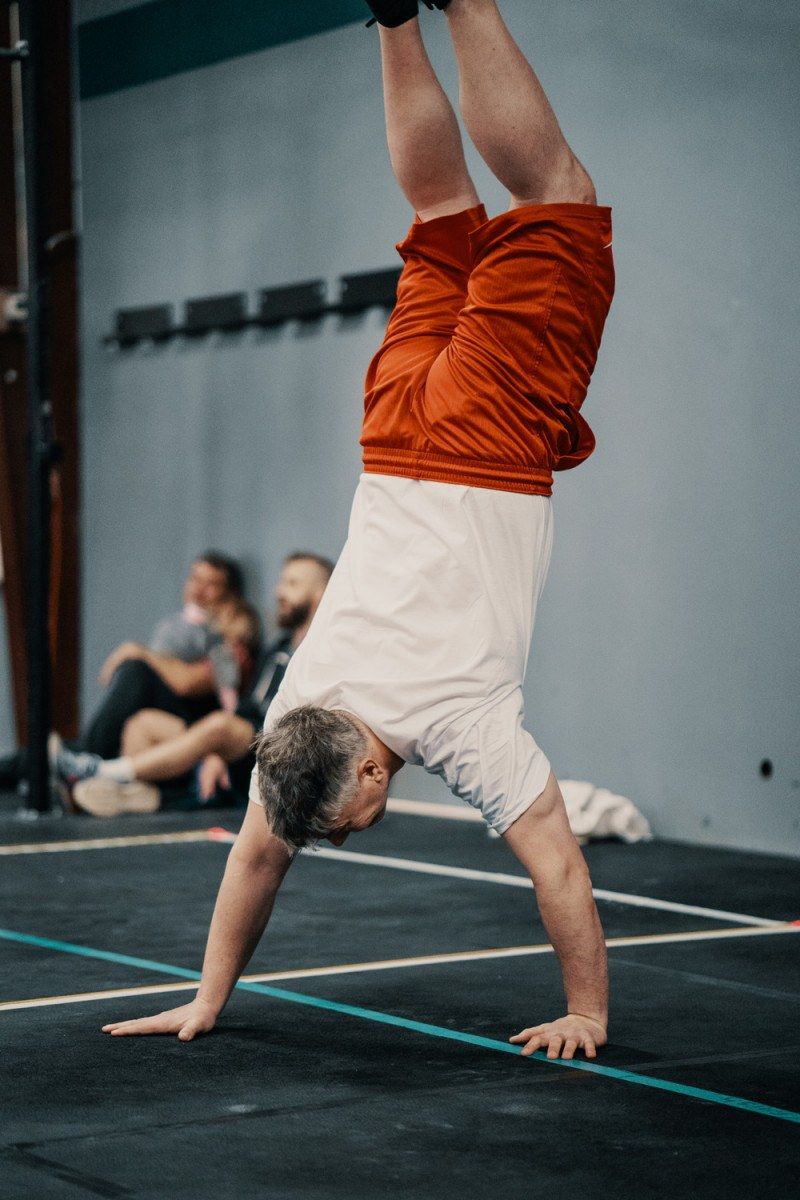 Person doing a handstand on a black floor in a gym, wearing white shirt and red shorts.