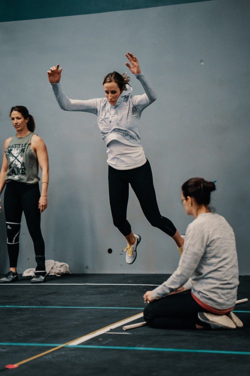 Woman jumping over a ball in a gym; another woman watches, two others stand nearby.