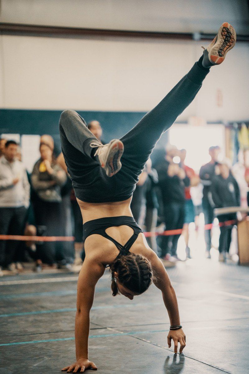 Woman in handstand, one leg raised, in a gym. Crowd watches in background.