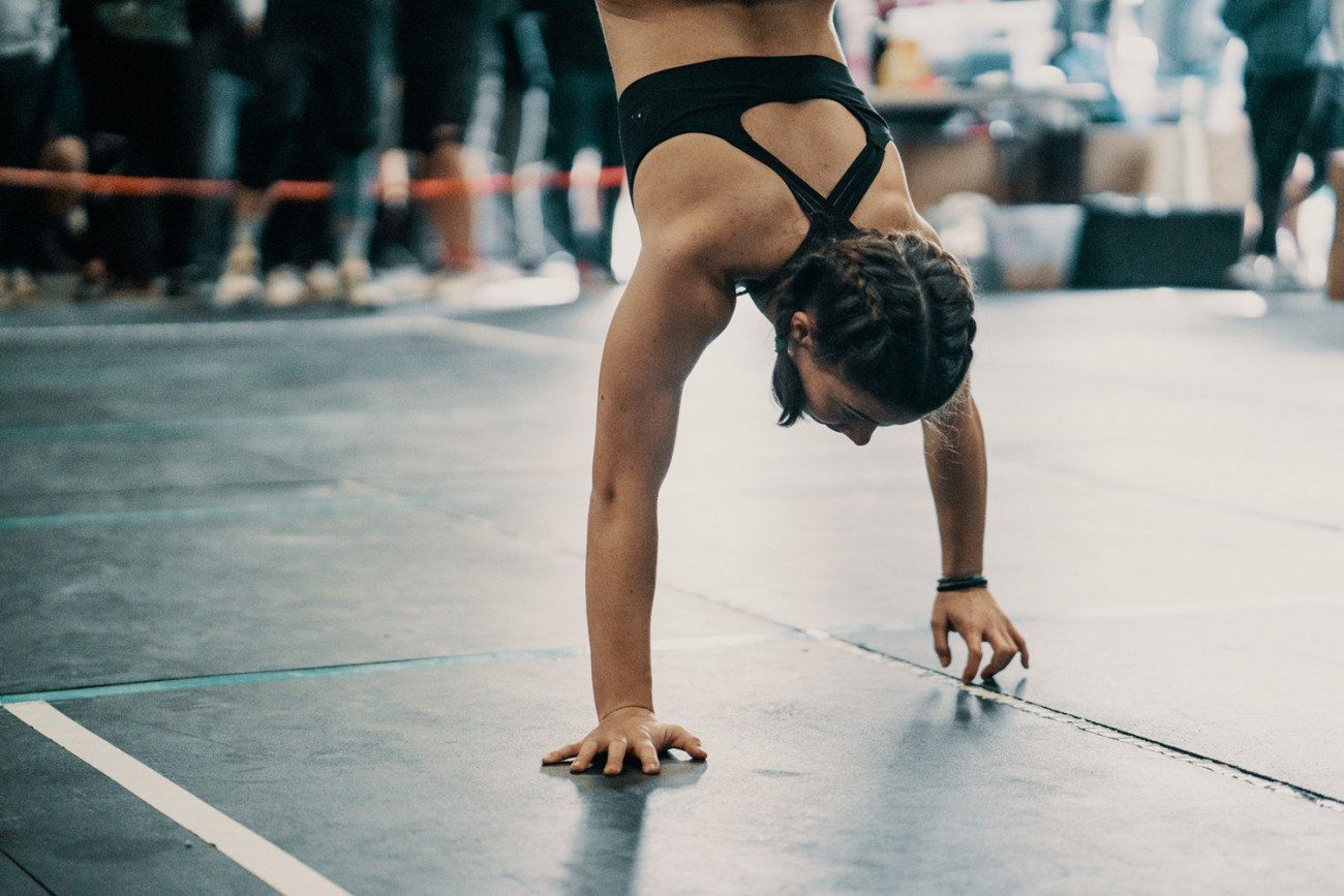 Woman doing a handstand on a grey floor, wearing black athletic wear. Braided hair. Crowd in background.