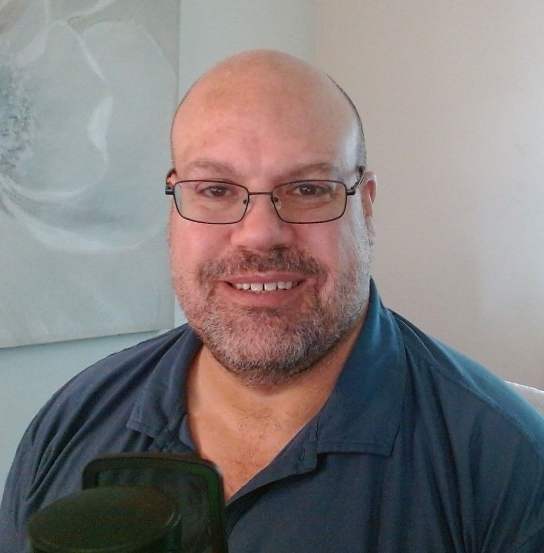 Man with glasses, bald head, and beard smiles at the camera, wearing a blue shirt.