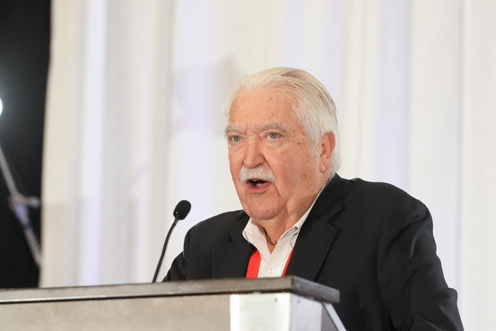 Man speaking at a podium, wearing a black jacket and white shirt. White hair and mustache, light background.