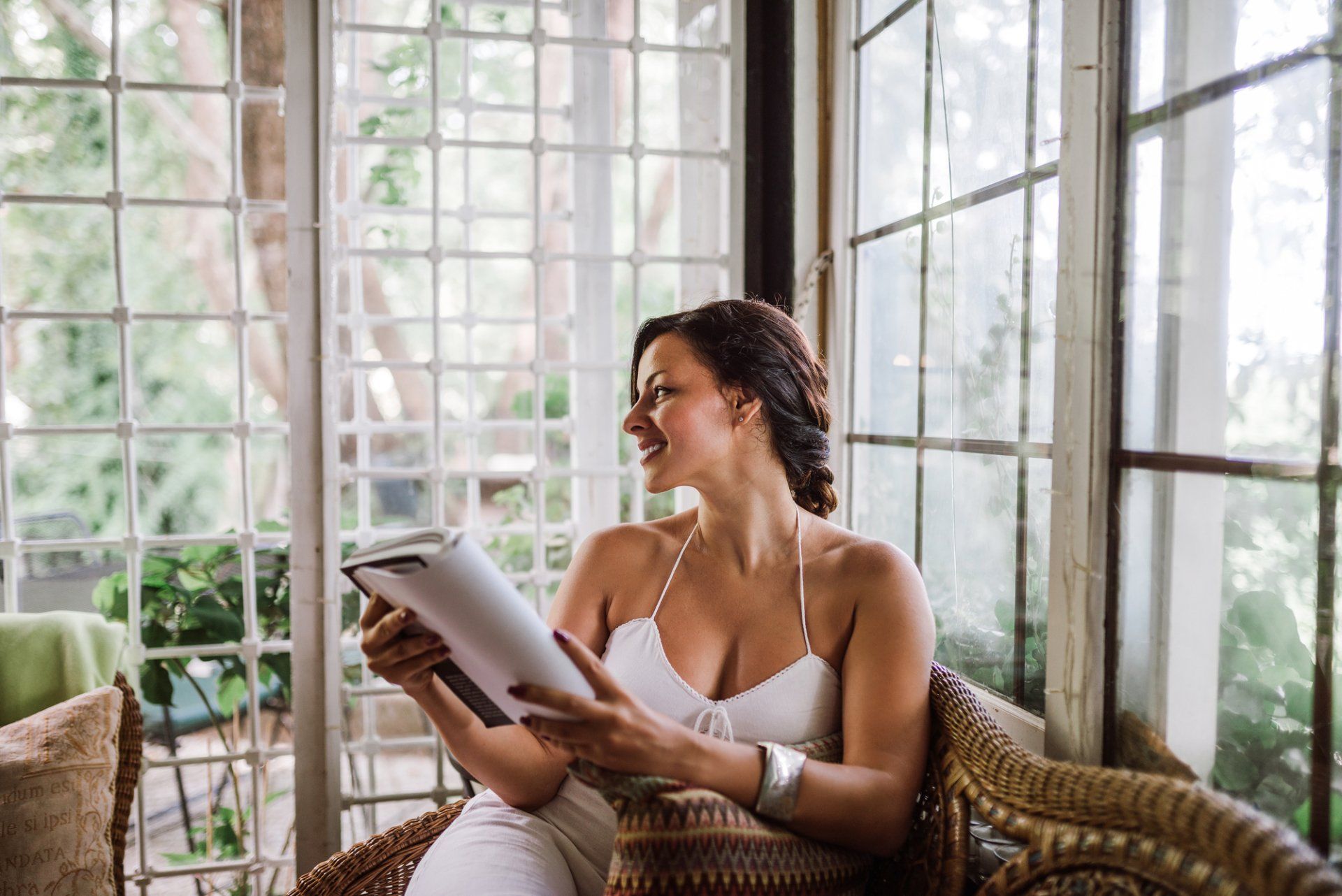 A woman is sitting in a wicker chair reading a book.