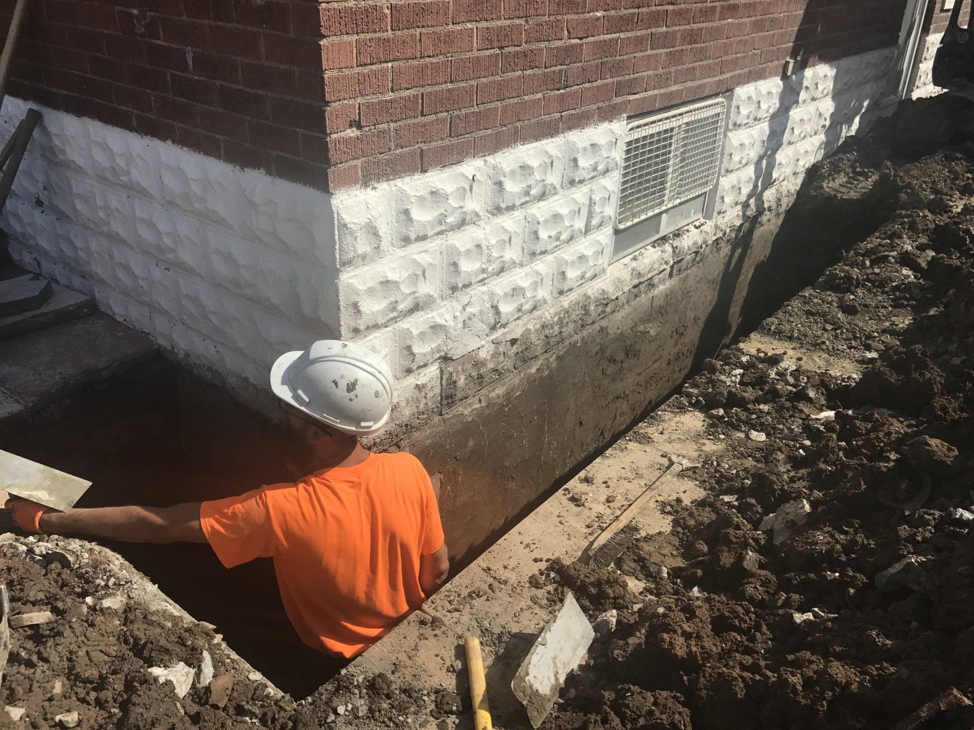 A man wearing a hard hat is digging a hole in front of a brick building.