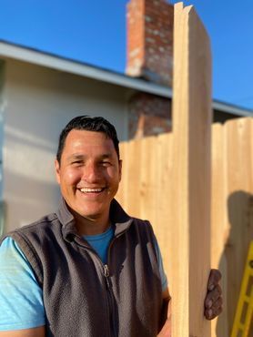 A man is smiling while holding a piece of wood in front of a wooden fence.
