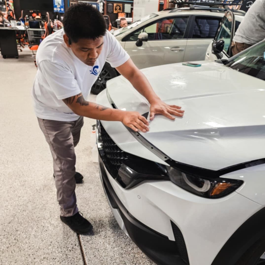 Man inspecting the hood of a white car indoors. He's wearing a white shirt, gray pants, and black shoes.
