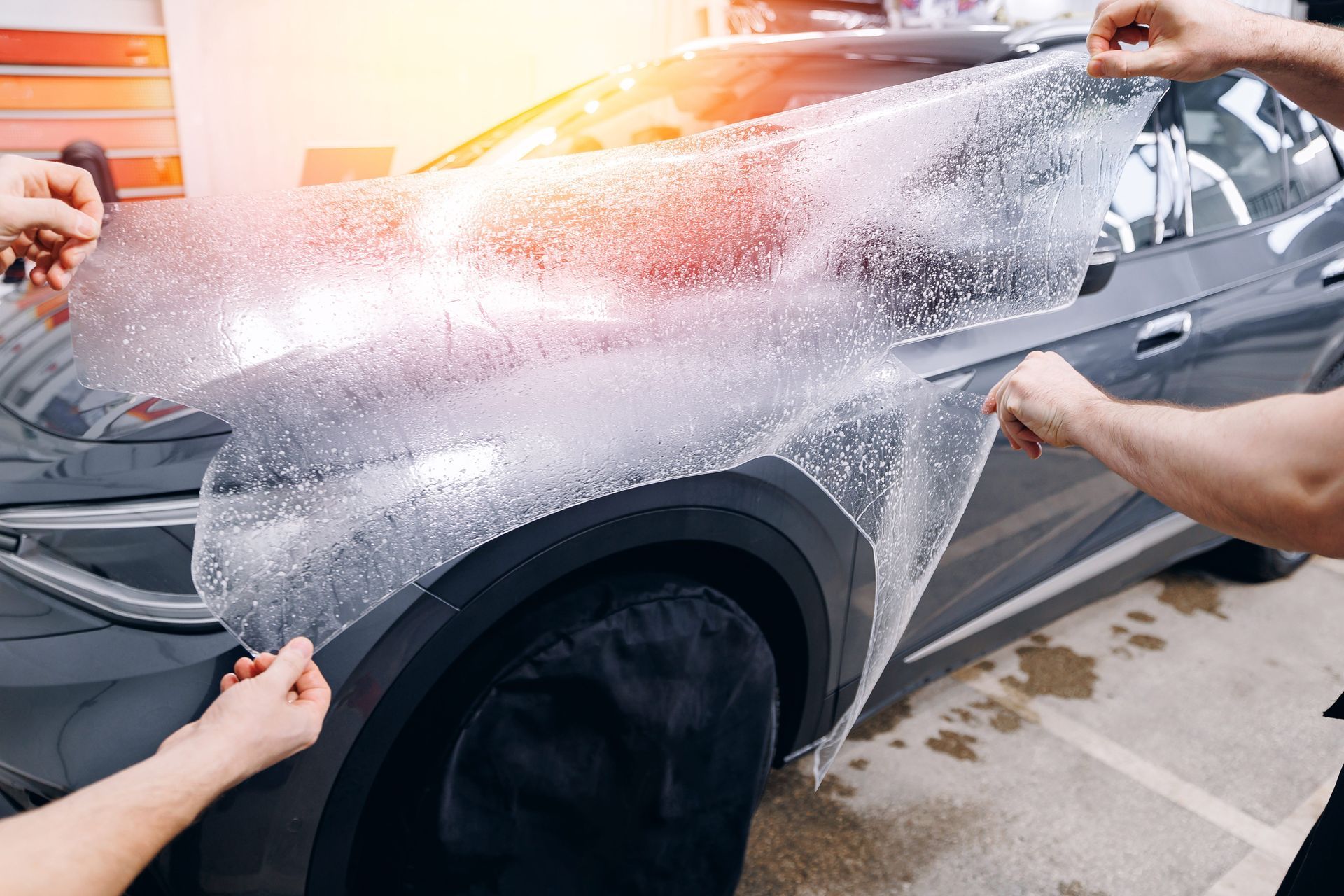 People applying clear paint protection film to the front quarter panel of a gray car.