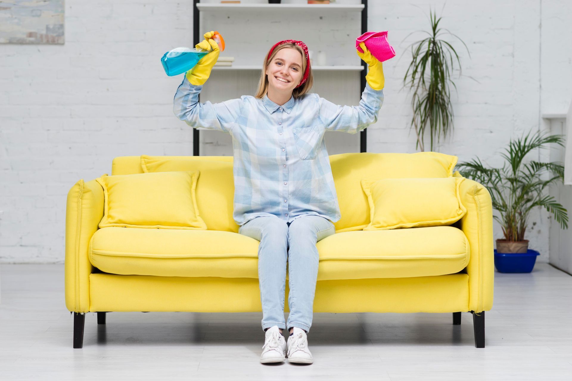 A woman is sitting on a yellow couch holding cleaning supplies.