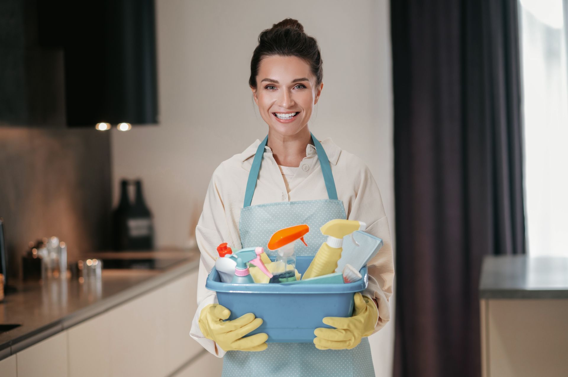 A woman is holding a bucket of cleaning supplies in a kitchen.