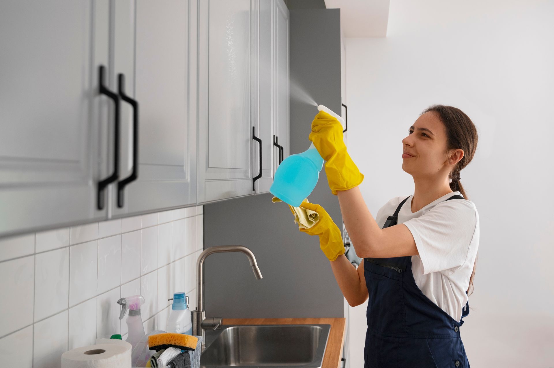 A woman is cleaning cabinets in a kitchen with a spray bottle.