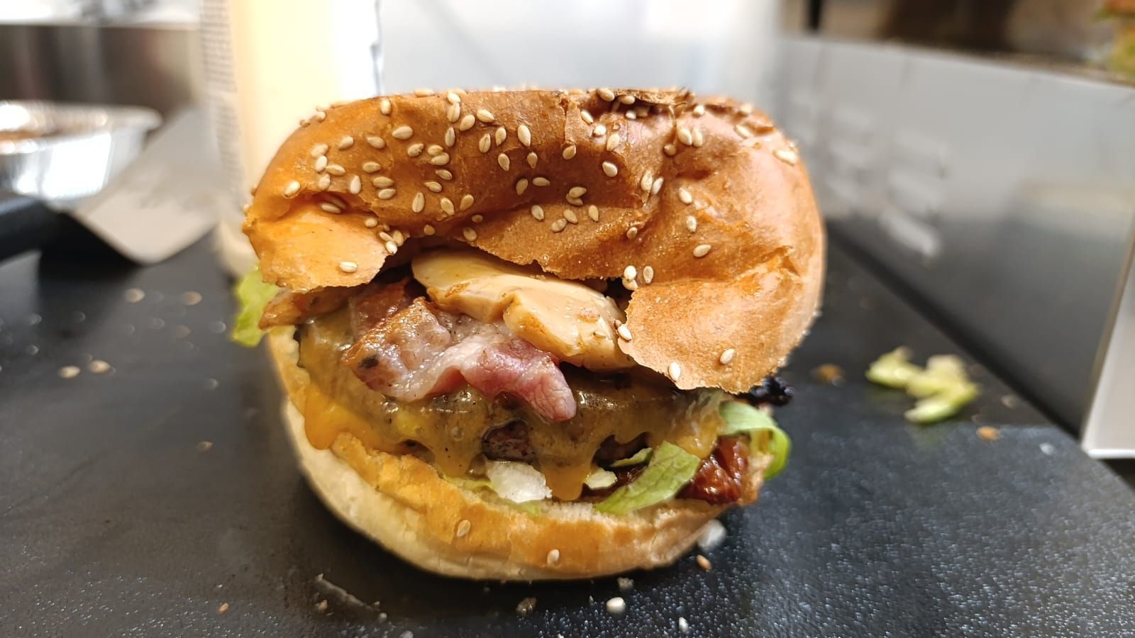 Hamburger con panino ai semi di sesamo, pancetta, lattuga e salsa su una superficie scura.