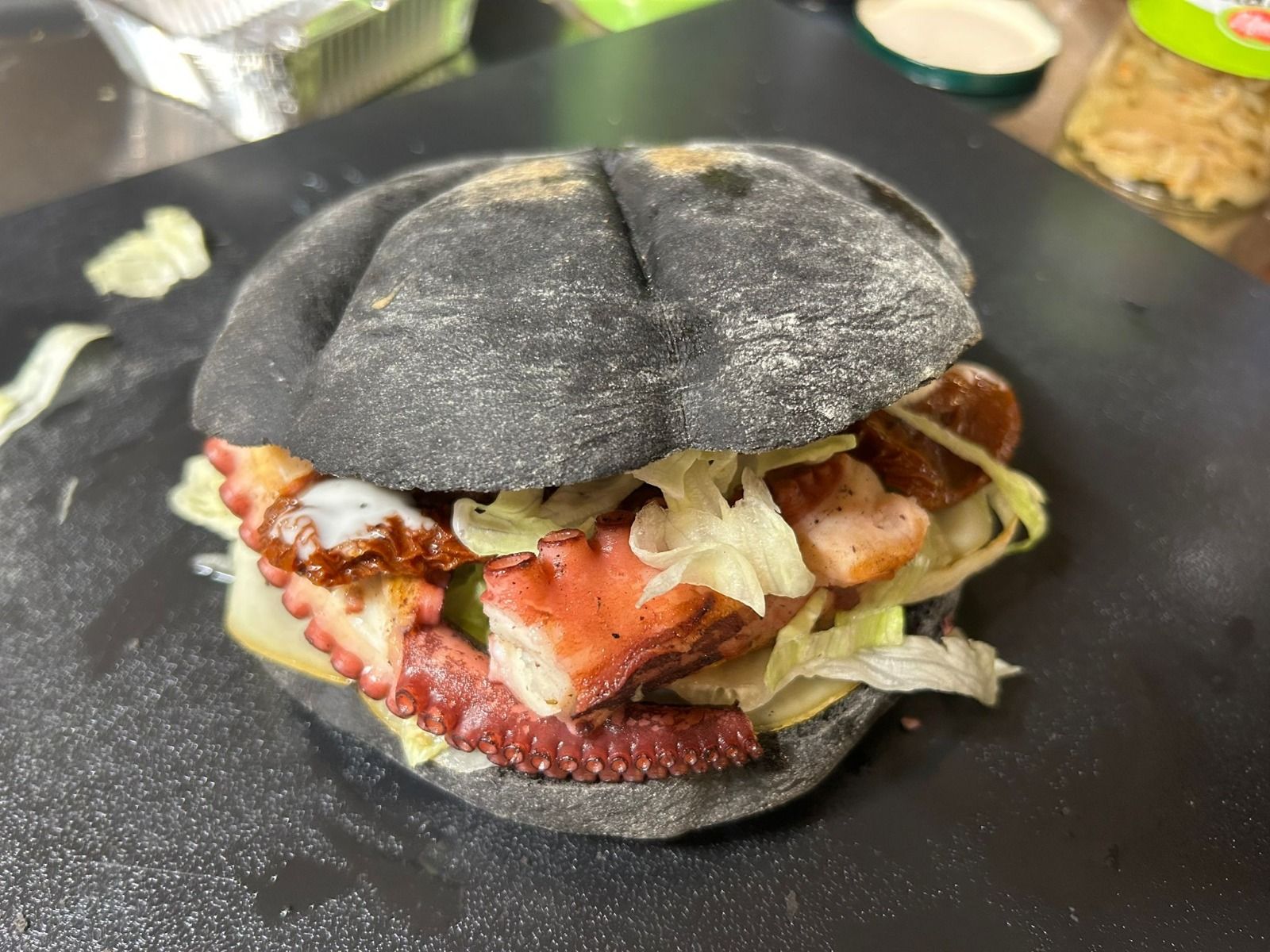 Panino nero con polpo, lattuga e salsa su un piatto nero.