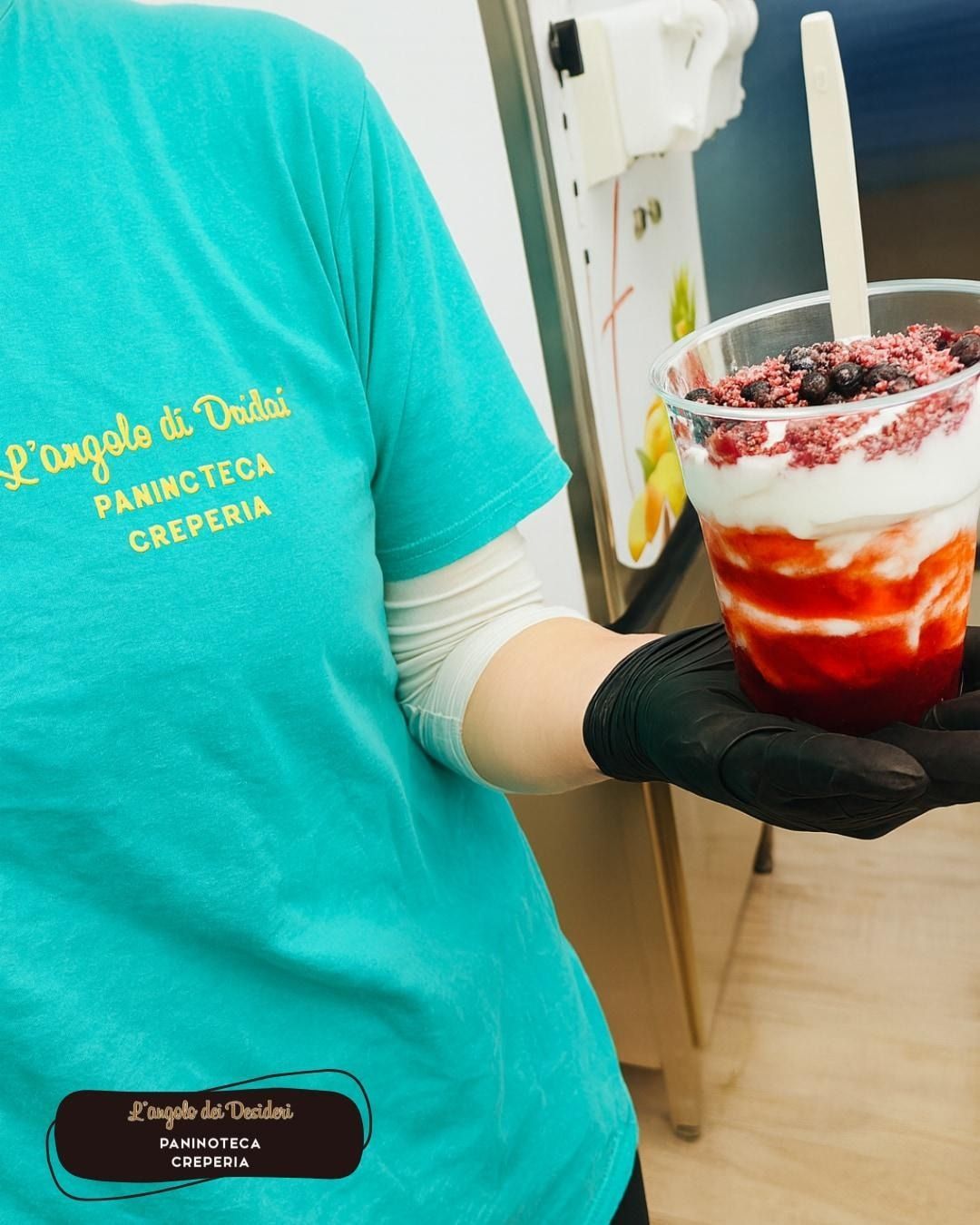 Persona che tiene in mano un parfait, con indosso una camicia verde acqua. Il parfait è composto da strati di salsa rossa, crema bianca e frutti di bosco.