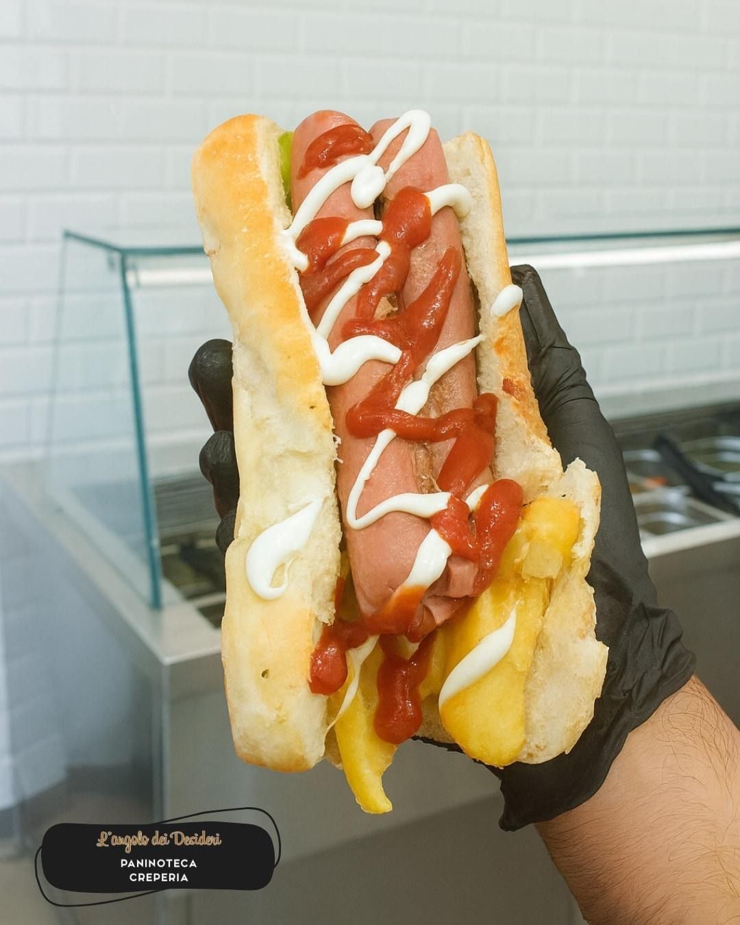 Hot dog in un panino, condito con ketchup, maionese e patatine fritte, tenuto da una mano guantata.