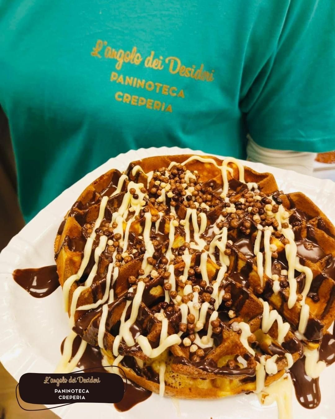 Waffle ricoperto di salsa al cioccolato, glassa bianca e granella, servito su un piatto di carta, tenuto in mano da una persona che indossa una maglietta verde.
