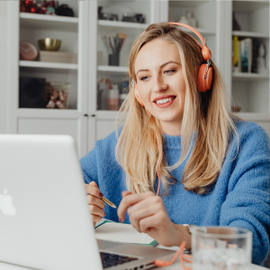 Smiling woman in blue sweater using a laptop at a desk, wearing orange headphones