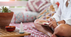Two women talking on a pink couch in a bright living room, one holding a notebook.