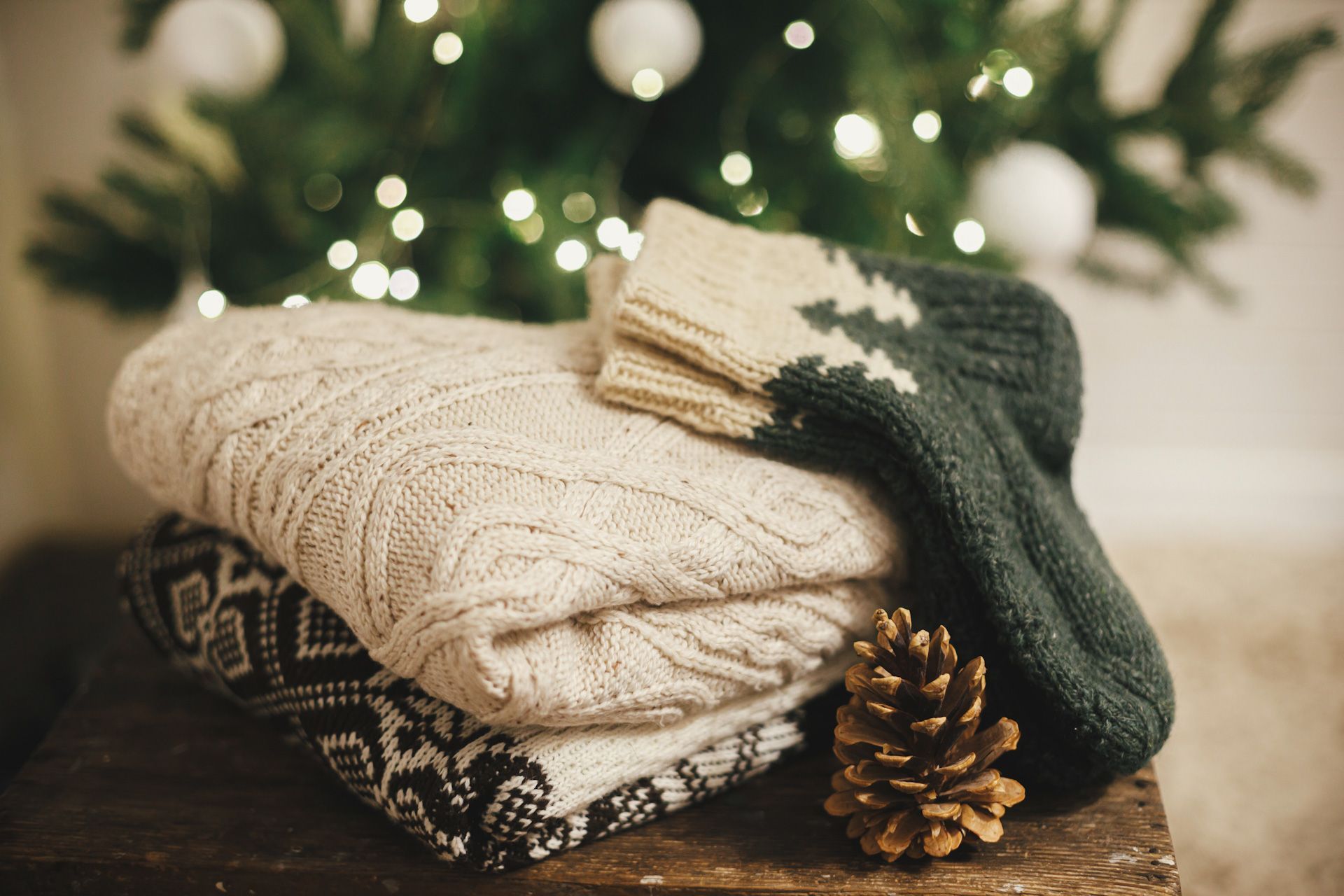 A stack of sweaters and a pine cone on a wooden table in front of a christmas tree .